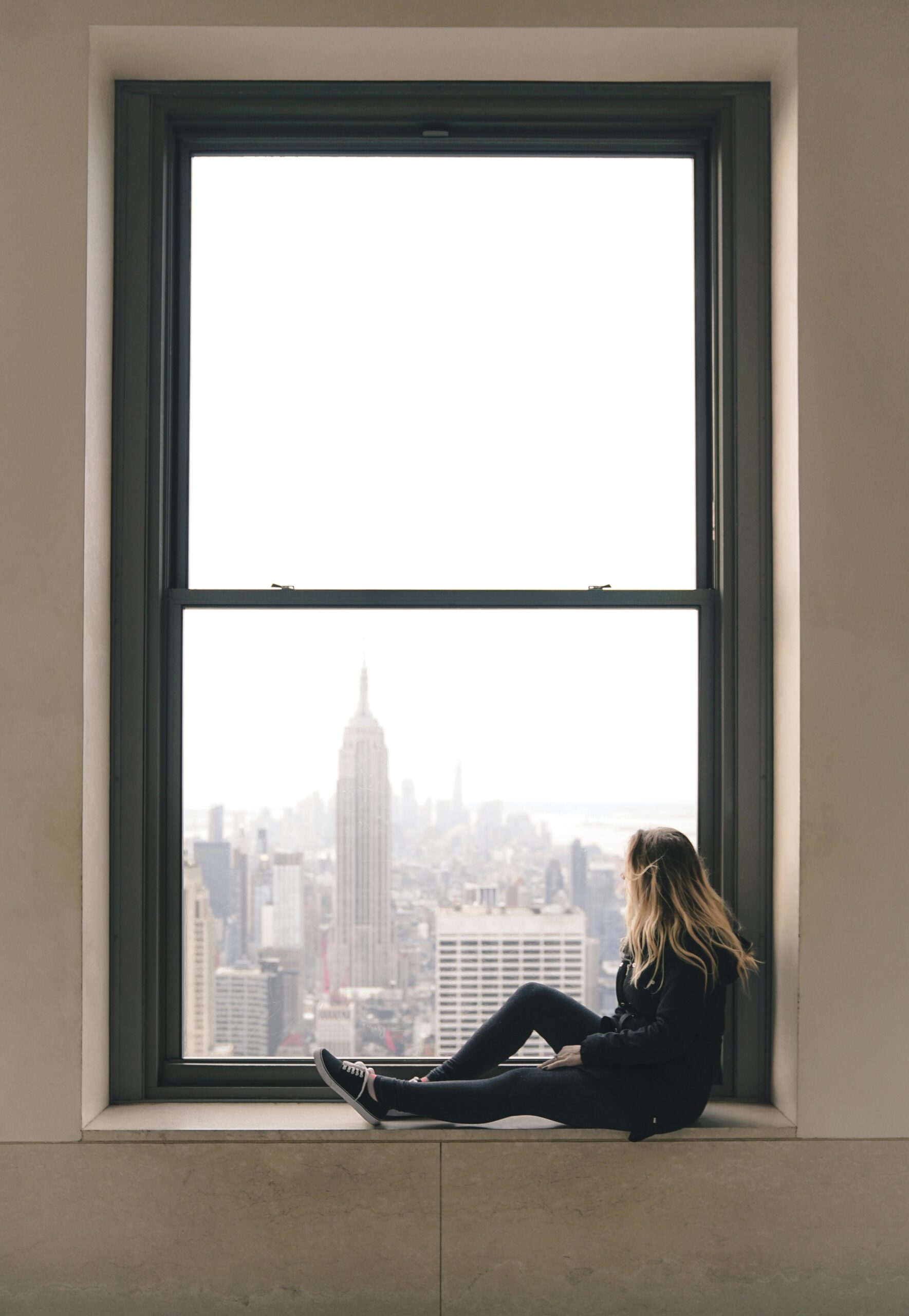 A woman sitting on the ledge of her New York City apartment window, gazing out thoughtfully at the skyline, reflecting on her life.